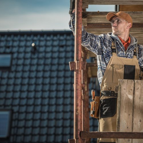 Covering House with Black Ceramic Roof Tiles. Caucasian Contractor in His 30s on Scaffolding.